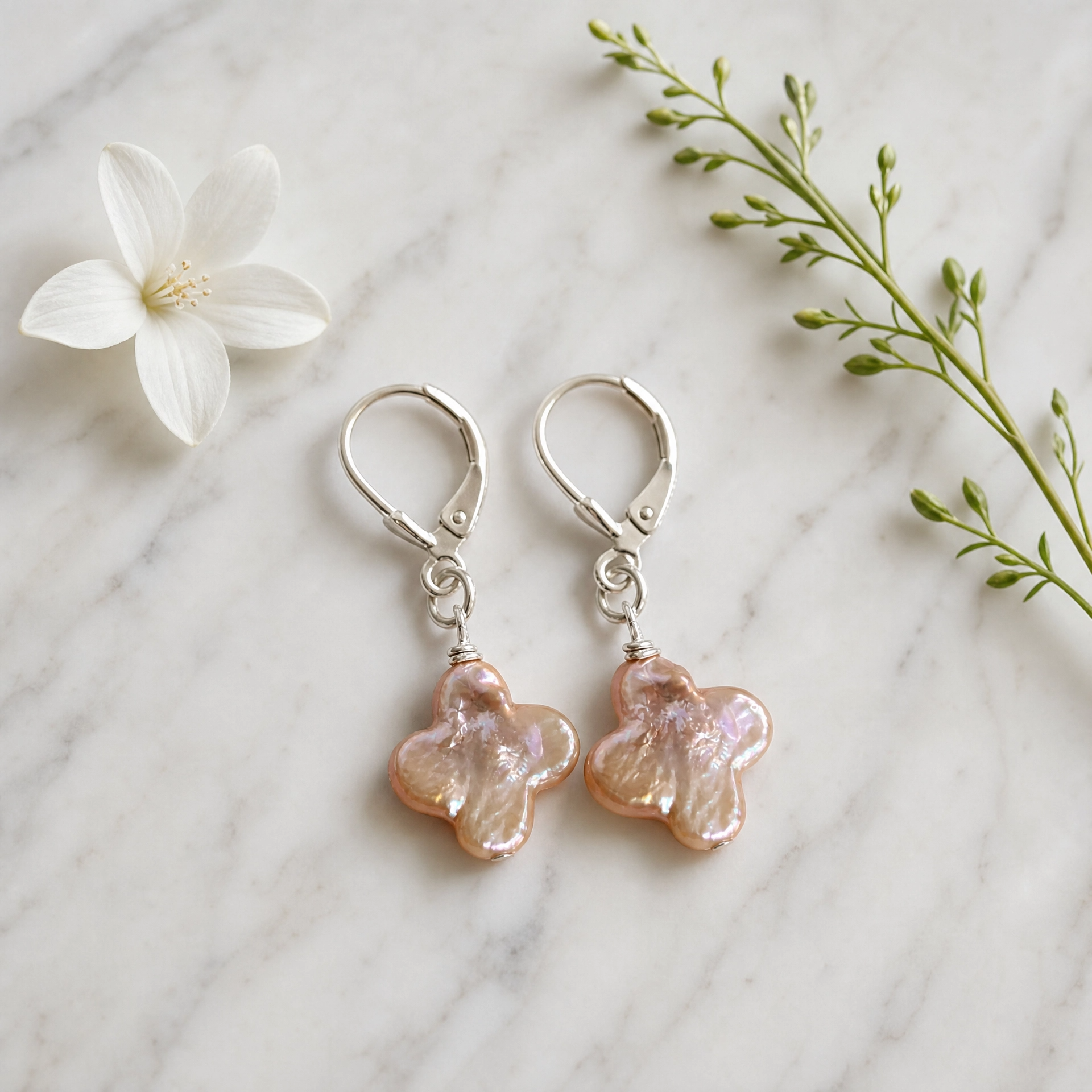 A pair of dangling earrings with silver leverback hooks and iridescent peach-pink flower-shaped charms, arranged on a white marble surface with a white flower and green sprig as props.