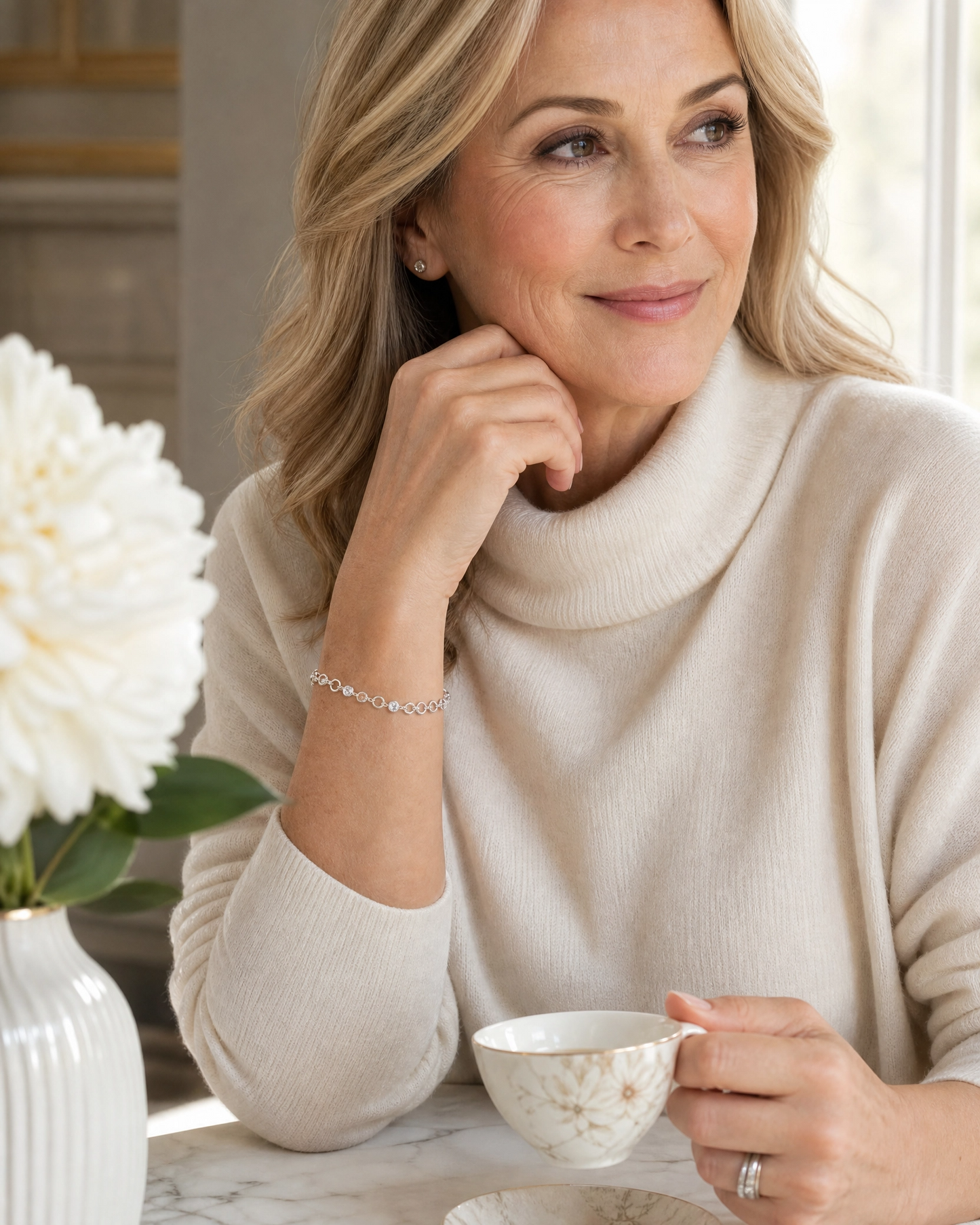 A woman with blonde hair wearing a cream-colored ribbed turtleneck sweater, holding a white teacup on a marble table with a white flower vase in the foreground.