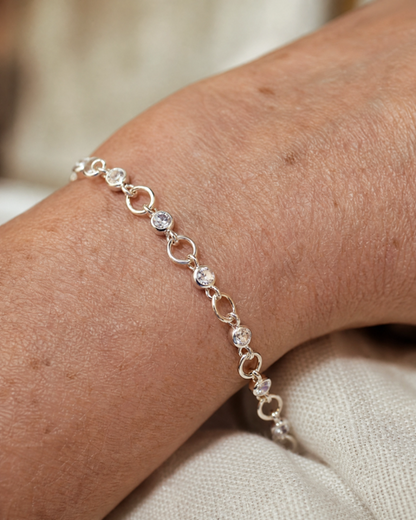 A delicate silver bracelet with small circular links and clear round stones, shown worn on a person's wrist against a soft beige fabric background.