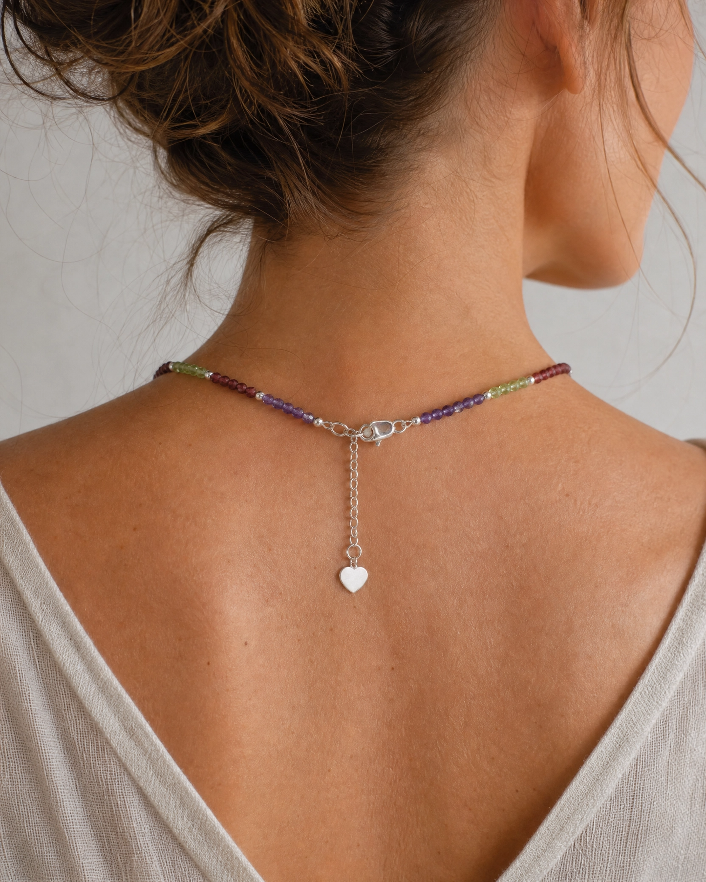A close-up back view of a woman wearing a delicate beaded necklace with a silver clasp and a small dangling heart charm.