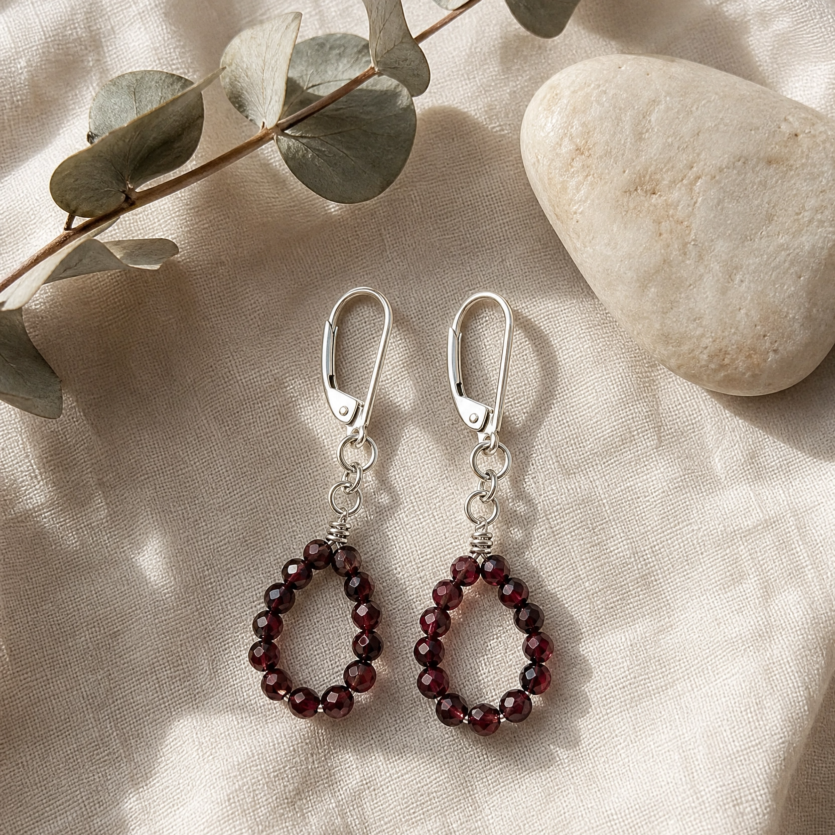 A pair of silver leverback drop earrings with dark red beaded teardrop shapes, displayed on a beige linen fabric alongside a stone and eucalyptus leaves.