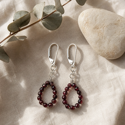 A pair of silver leverback drop earrings with dark red beaded teardrop shapes, displayed on a beige linen fabric alongside a stone and eucalyptus leaves.