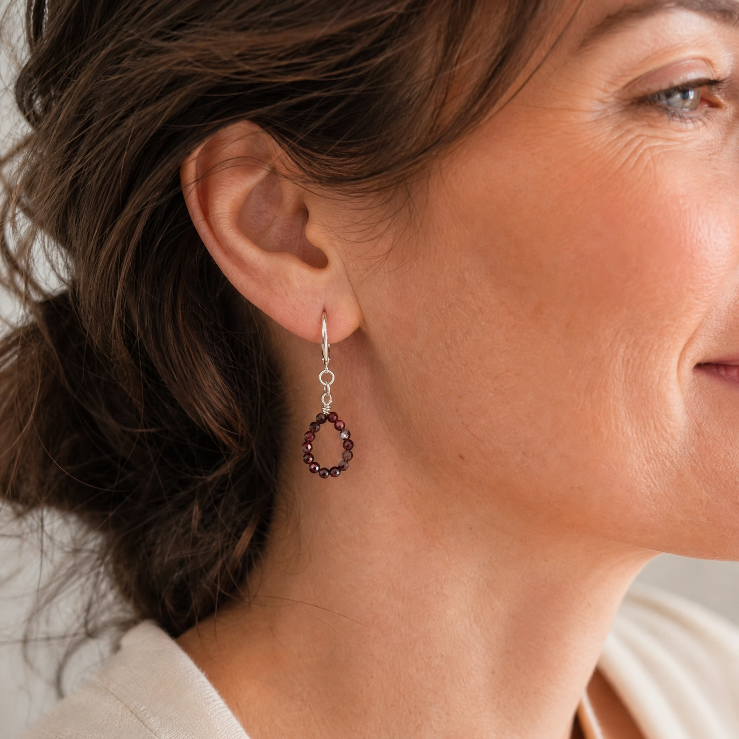 A close-up of a woman wearing a delicate silver-toned dangle earring with a small circular cluster of dark red gemstones.