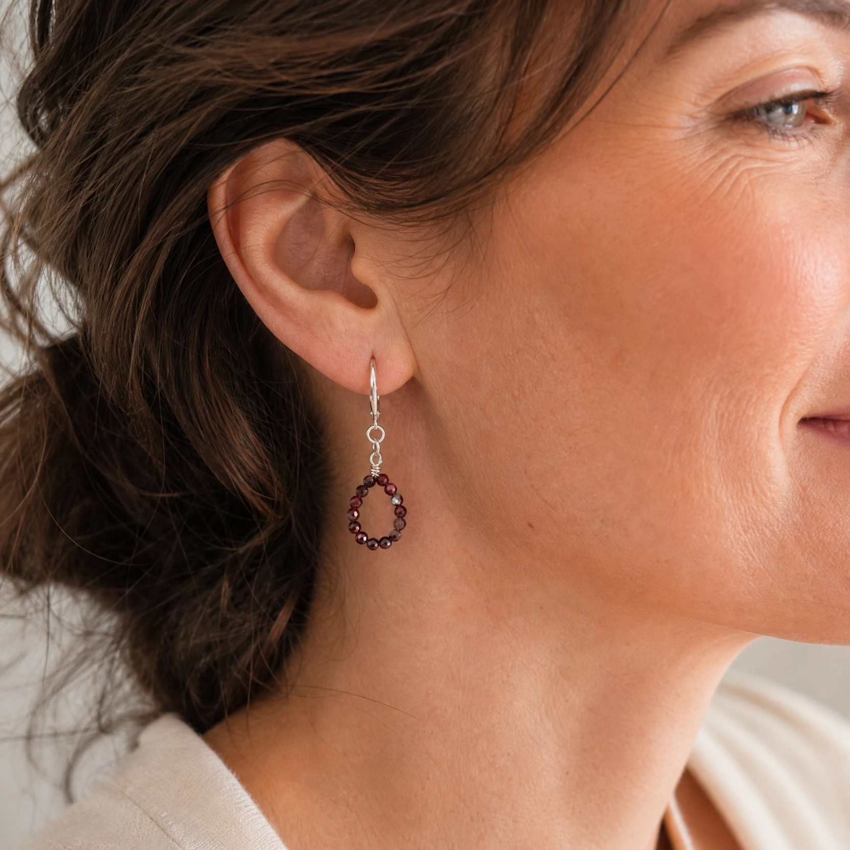 A close-up of a woman wearing a delicate silver-toned dangle earring with a small circular cluster of dark red gemstones.