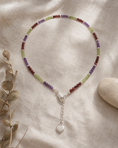 A beaded necklace with alternating purple, green, and red beads, finished with a silver lobster clasp and a dangling silver heart charm, displayed on a textured beige linen fabric with dried eucalyptus leaves and a stone.