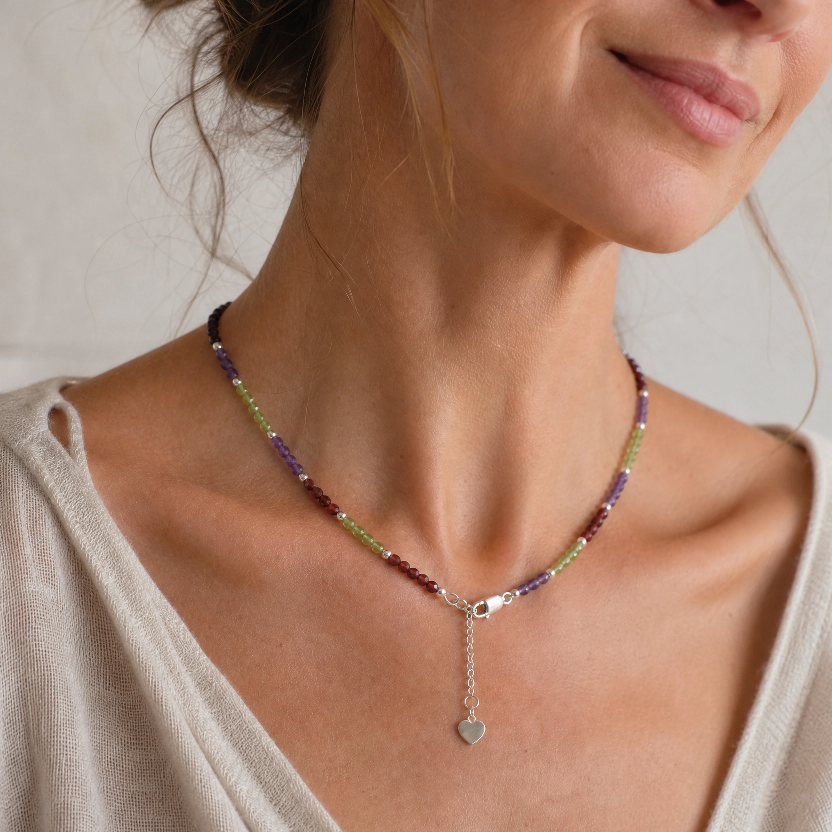 A close-up of a woman wearing a delicate beaded necklace with a silver clasp and a small dangling heart charm. The necklace features small beads in purple, green, and red tones.