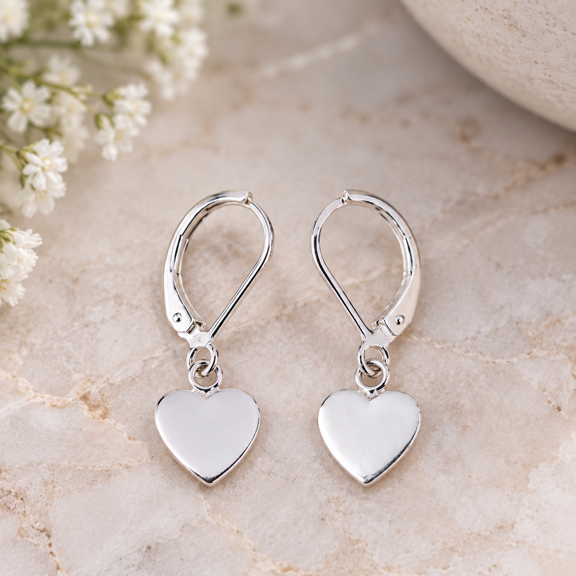 A pair of polished silver leverback earrings with flat heart-shaped dangles, displayed on a light beige marble surface with small white flowers in the background.