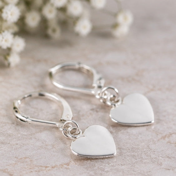 A pair of silver leverback earrings with smooth, polished heart-shaped dangles, displayed on a light beige textured surface with blurred white baby's breath flowers in the background.