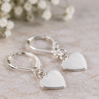 A pair of silver leverback earrings with smooth, polished heart-shaped dangles, displayed on a light beige textured surface with blurred white baby's breath flowers in the background.