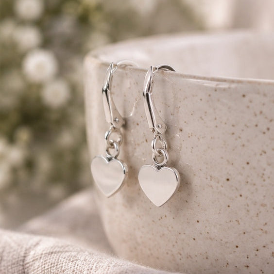 A pair of silver heart dangle earrings resting on the rim of a speckled ceramic mug, with a soft-focus floral background and textured fabric in the foreground.