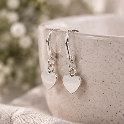 A pair of silver heart dangle earrings resting on the rim of a speckled ceramic mug, with a soft-focus floral background and textured fabric in the foreground.