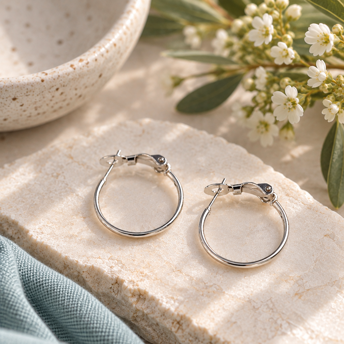 A pair of polished silver hoop earrings displayed on a textured beige stone slab, styled with a light blue fabric, white flowers, and a ceramic bowl in soft natural light.