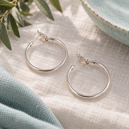 A pair of polished silver hoop earrings displayed on a textured beige linen cloth, styled with a light blue ceramic bowl and green olive leaves.
