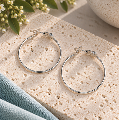 A pair of polished silver hoop earrings displayed on a textured beige stone slab, styled with a blue fabric napkin, small white flowers, and a ceramic bowl.
