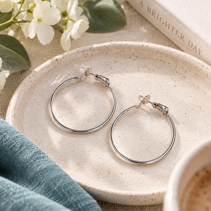 A pair of polished silver hoop earrings resting on a speckled beige ceramic dish, styled with white flowers, a blue textured cloth, a book, and a cup of coffee.