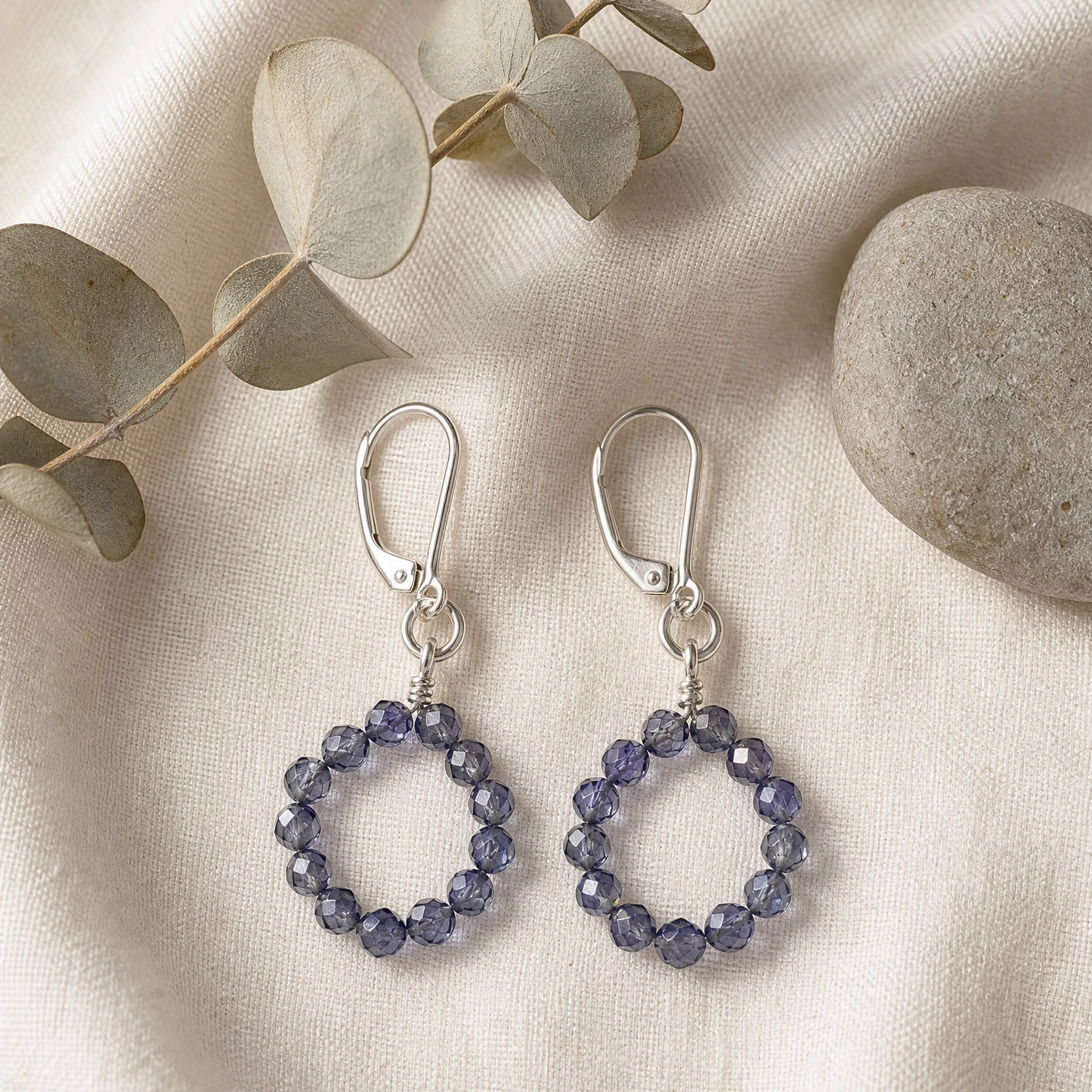 A pair of silver drop earrings with circular beaded rings in a periwinkle blue-purple color, displayed on a textured cream fabric with eucalyptus leaves and a stone.