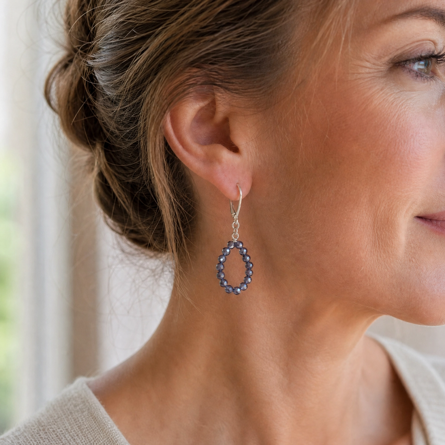 A close-up of a woman wearing a dangling hoop earring with a silver-tone frame and small dark blue stones, shown against a softly blurred indoor background.