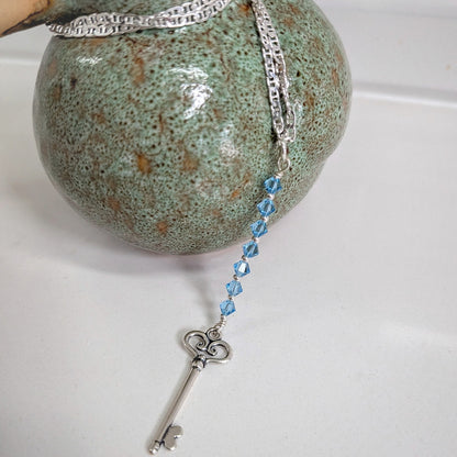 A silver key pendant with a strand of swarovski aquamarine crystals displayed on a white surface next to a green speckled ceramic object.