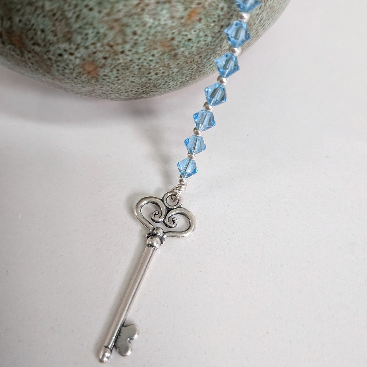 A silver key pendant with a series of aquamarine swarovski crystals, displayed on a white surface next to a textured green stone.