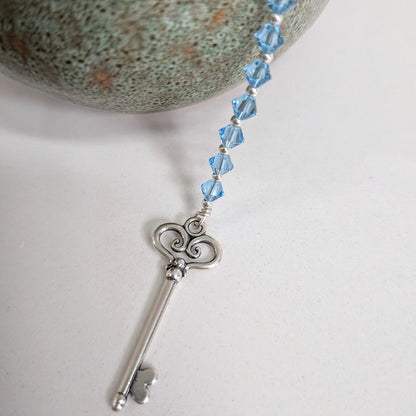 A silver key pendant with a series of aquamarine swarovski crystals, displayed on a white surface next to a textured green stone.