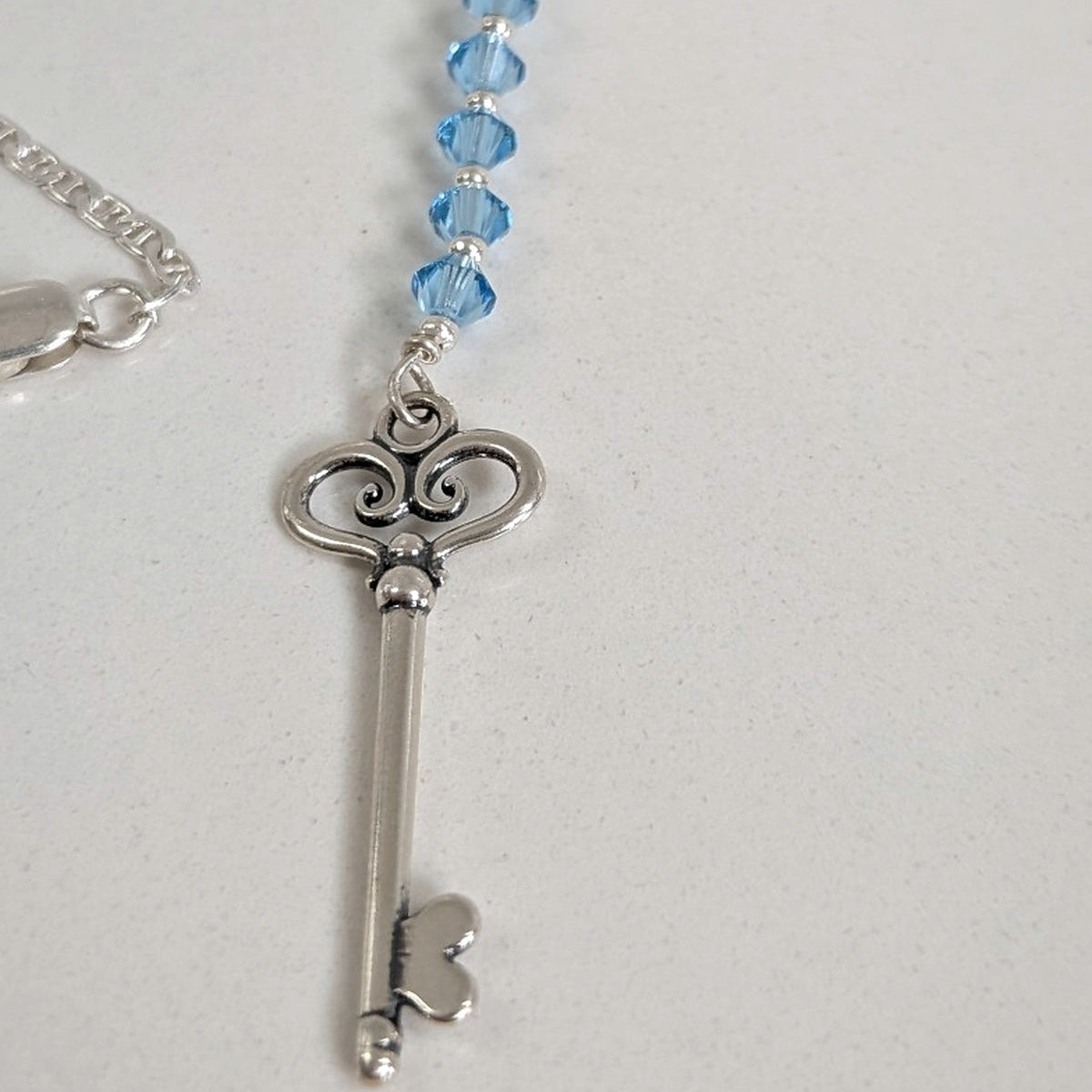 A silver key pendant charm necklace with a beaded top featuring swarovski aquamarine crystals, shown on a plain light gray background.