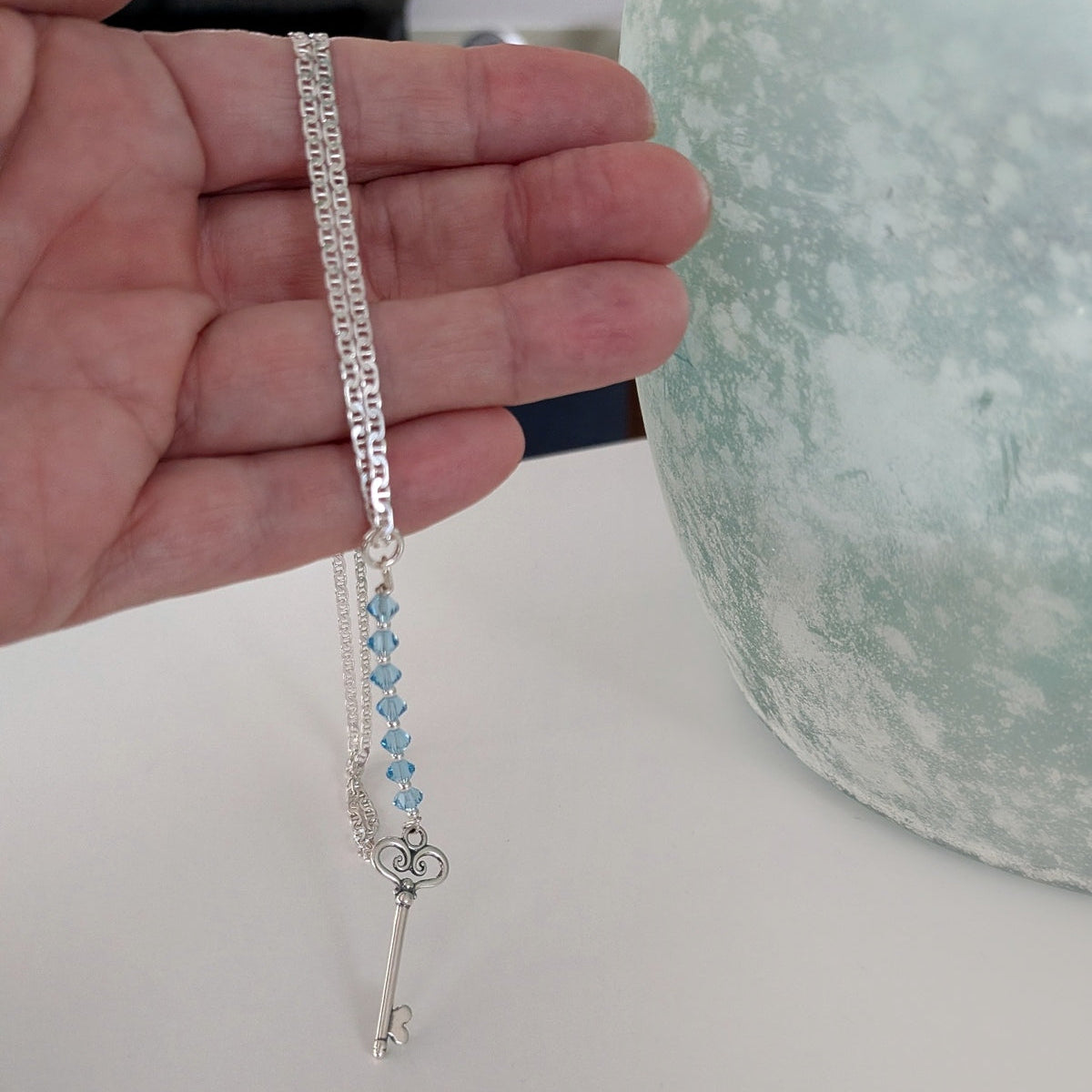 A silver key pendant necklace with a chain and swarovski aquamarine crystals, held in a person's hand against a white tabletop with a textured vase in the background.