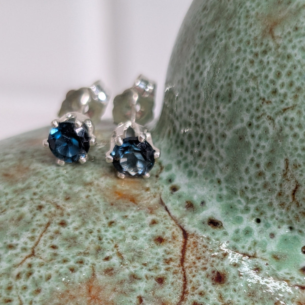 A close-up of a pair of silver stud earrings with deep blue gemstones, resting on a textured, glossy green ceramic surface.