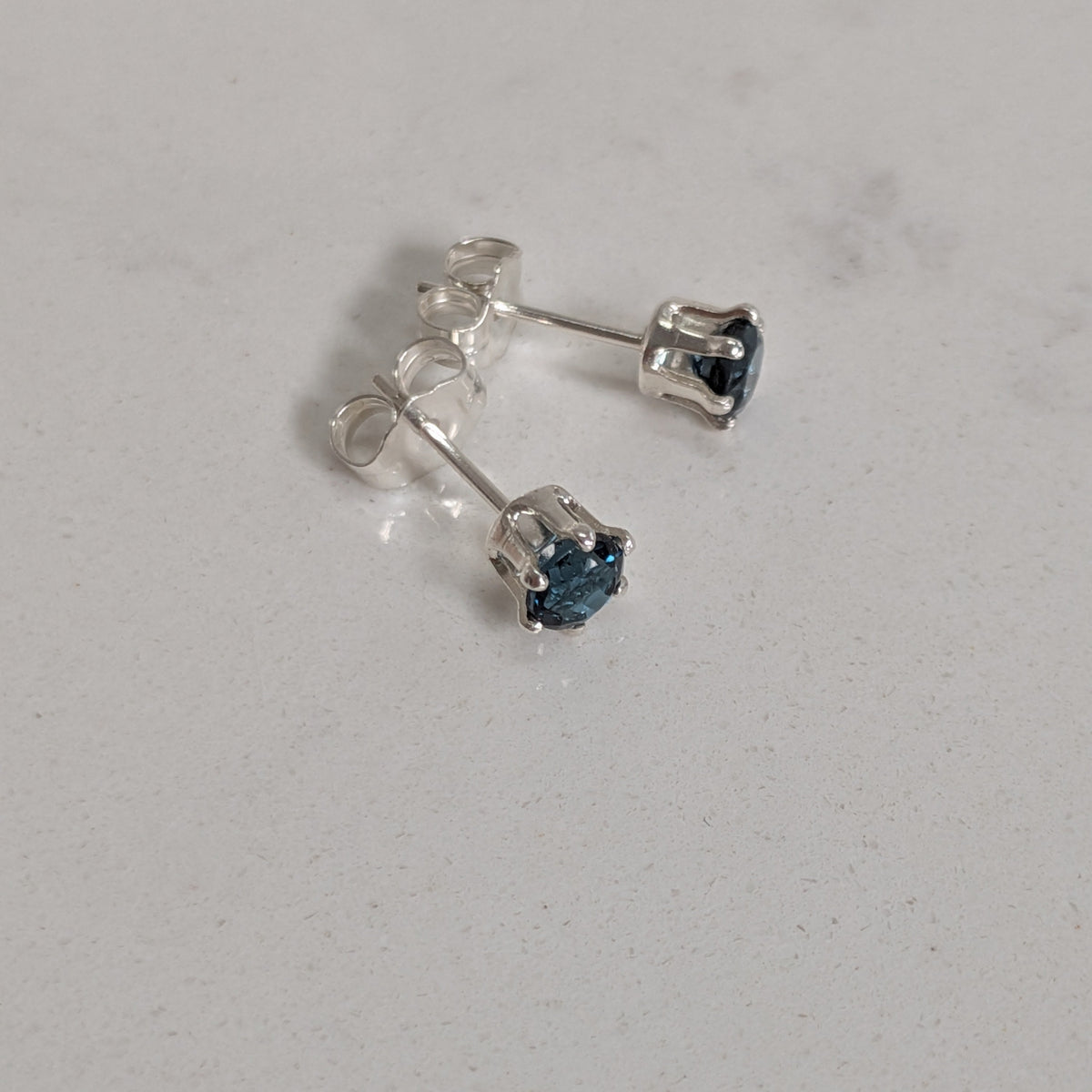 A pair of silver stud earrings with small dark blue heart-shaped gemstones, shown on a light gray textured surface.
