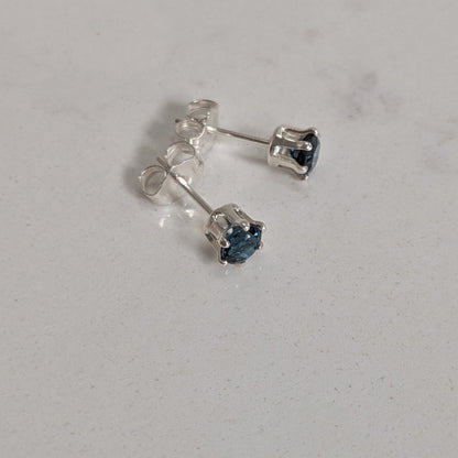 A pair of silver stud earrings with small dark blue heart-shaped gemstones, shown on a light gray textured surface.