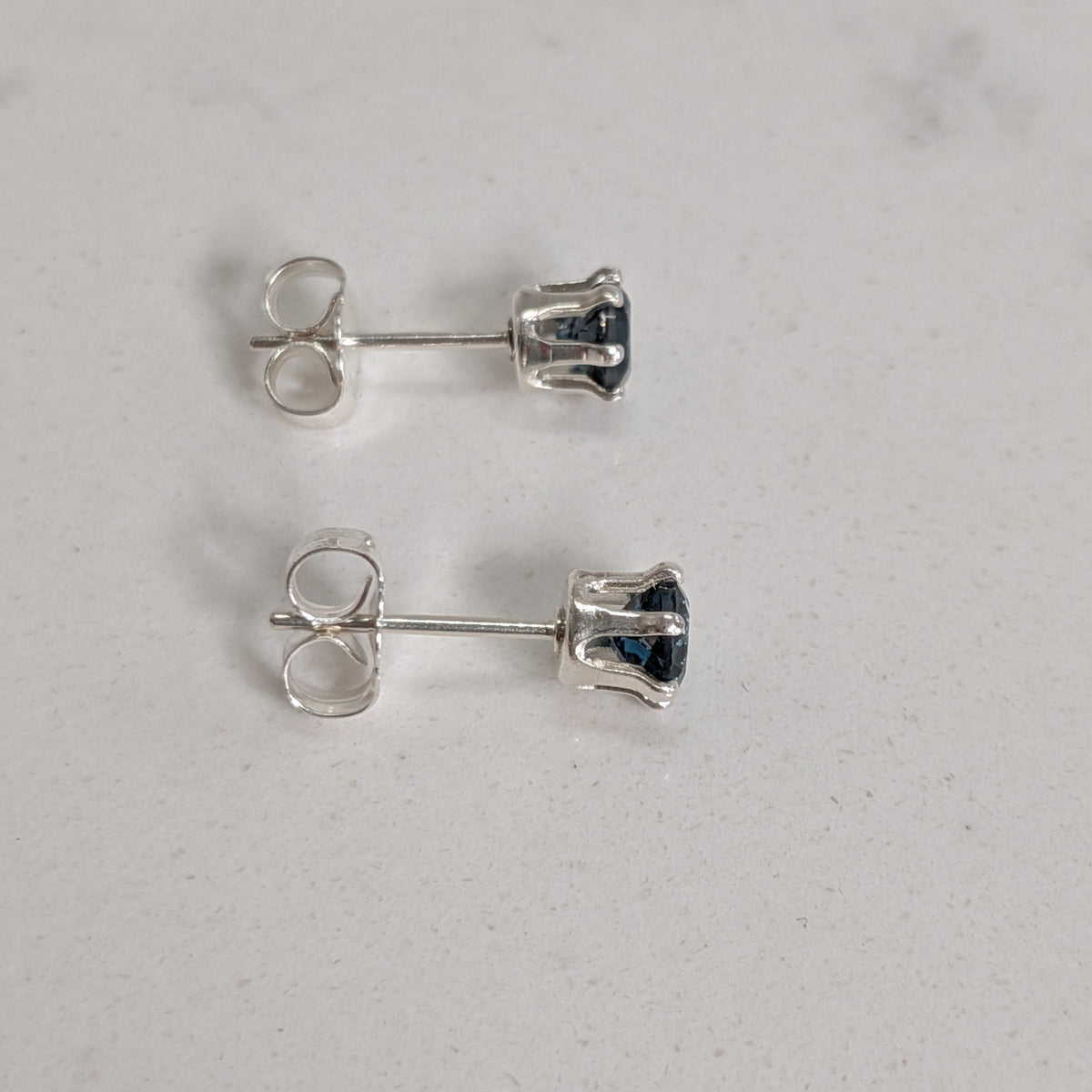 A pair of silver stud earrings with small dark blue gemstones, shown from the back on a light gray textured surface.