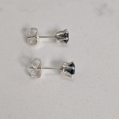 A pair of silver stud earrings with small dark blue gemstones, shown from the back on a light gray textured surface.