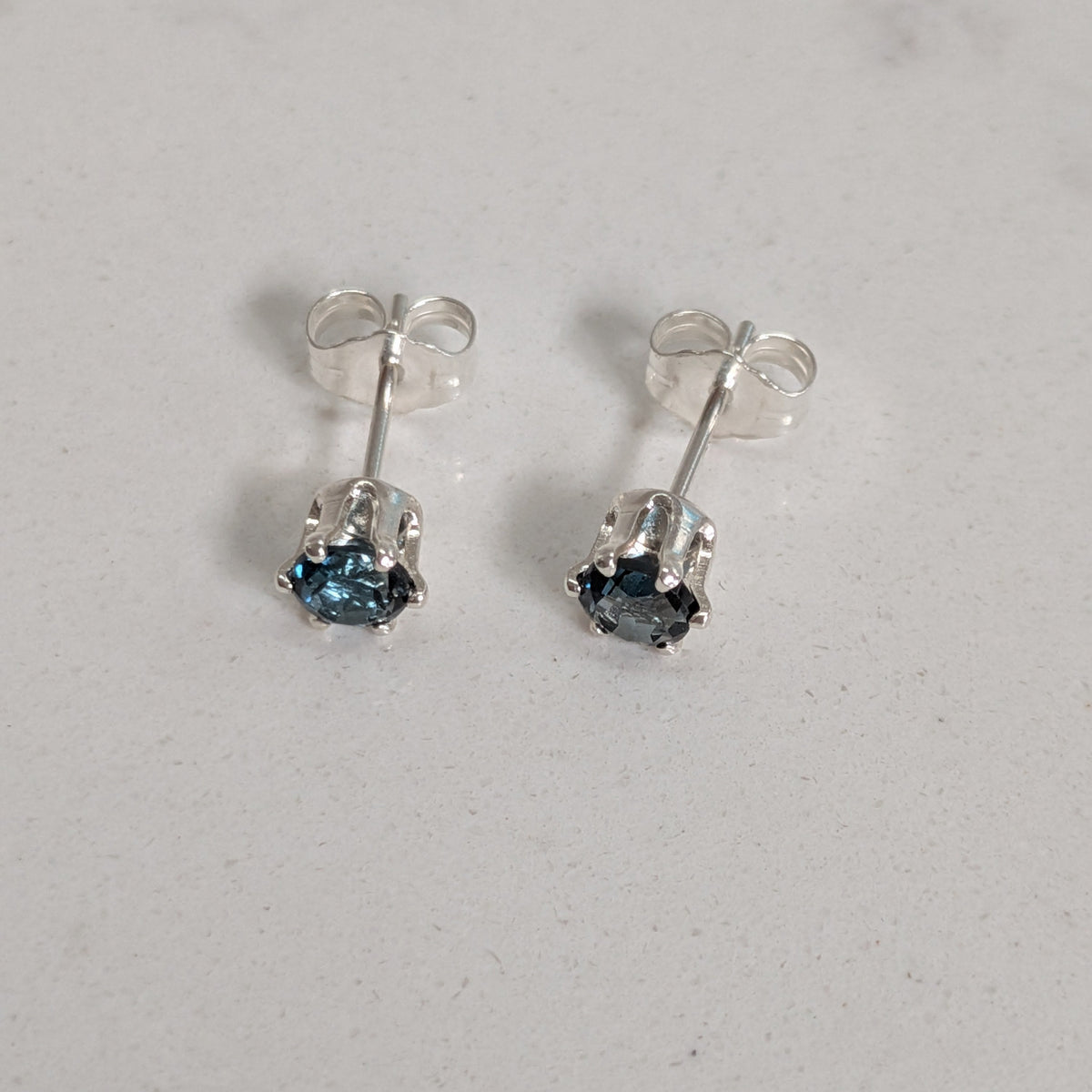 A pair of silver stud earrings with small round blue gemstones, shown on a light gray textured surface.