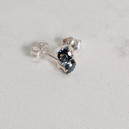 A pair of silver stud earrings with dark blue oval gemstones, shown on a light gray textured surface.