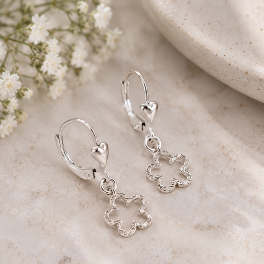 A pair of silver leverback earrings with textured flower charms and heart-shaped side details, displayed on a light marble surface with small white flowers and a ceramic dish in the background.