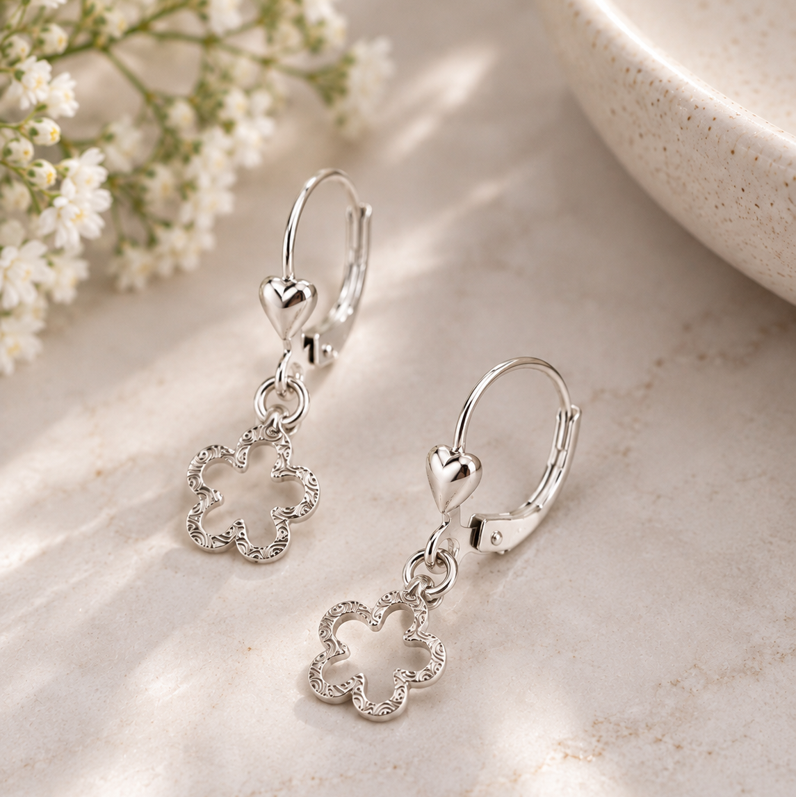 A pair of silver leverback earrings with small heart charms and dangling flower-shaped charms set with clear stones, displayed on a light marble surface with soft shadows and small white flowers.