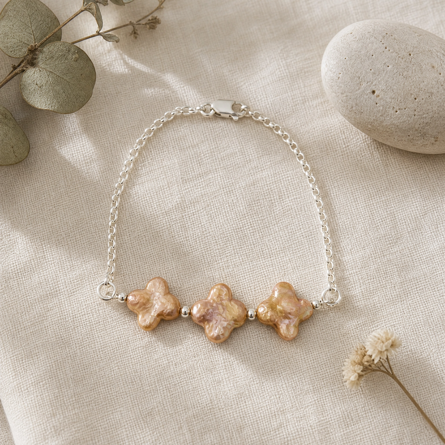 A delicate silver chain bracelet featuring three textured, champagne four leaf clover pearls, styled on a beige linen fabric with dried eucalyptus leaves and a stone.