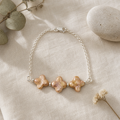A delicate silver chain bracelet featuring three textured, champagne four leaf clover pearls, styled on a beige linen fabric with dried eucalyptus leaves and a stone.