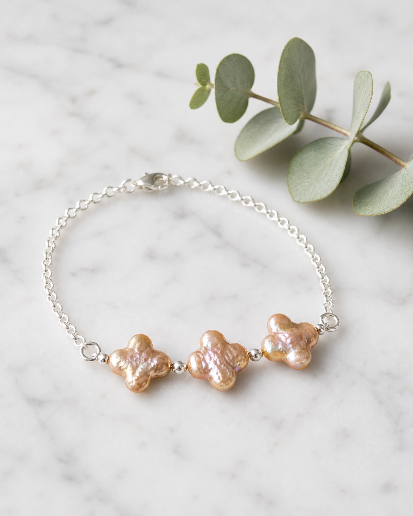 A delicate silver chain bracelet with three textured, champagne four leaf clover pearls, displayed on a white marble surface next to a green eucalyptus branch.
