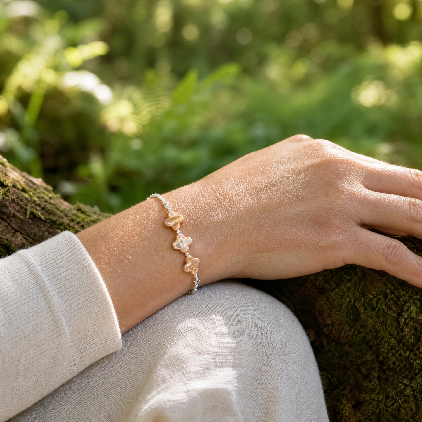 A delicate silver rolo chain bracelet with champagne four leaf clover pearls worn on a person's wrist, shown in a natural outdoor setting with a blurred green forest background.