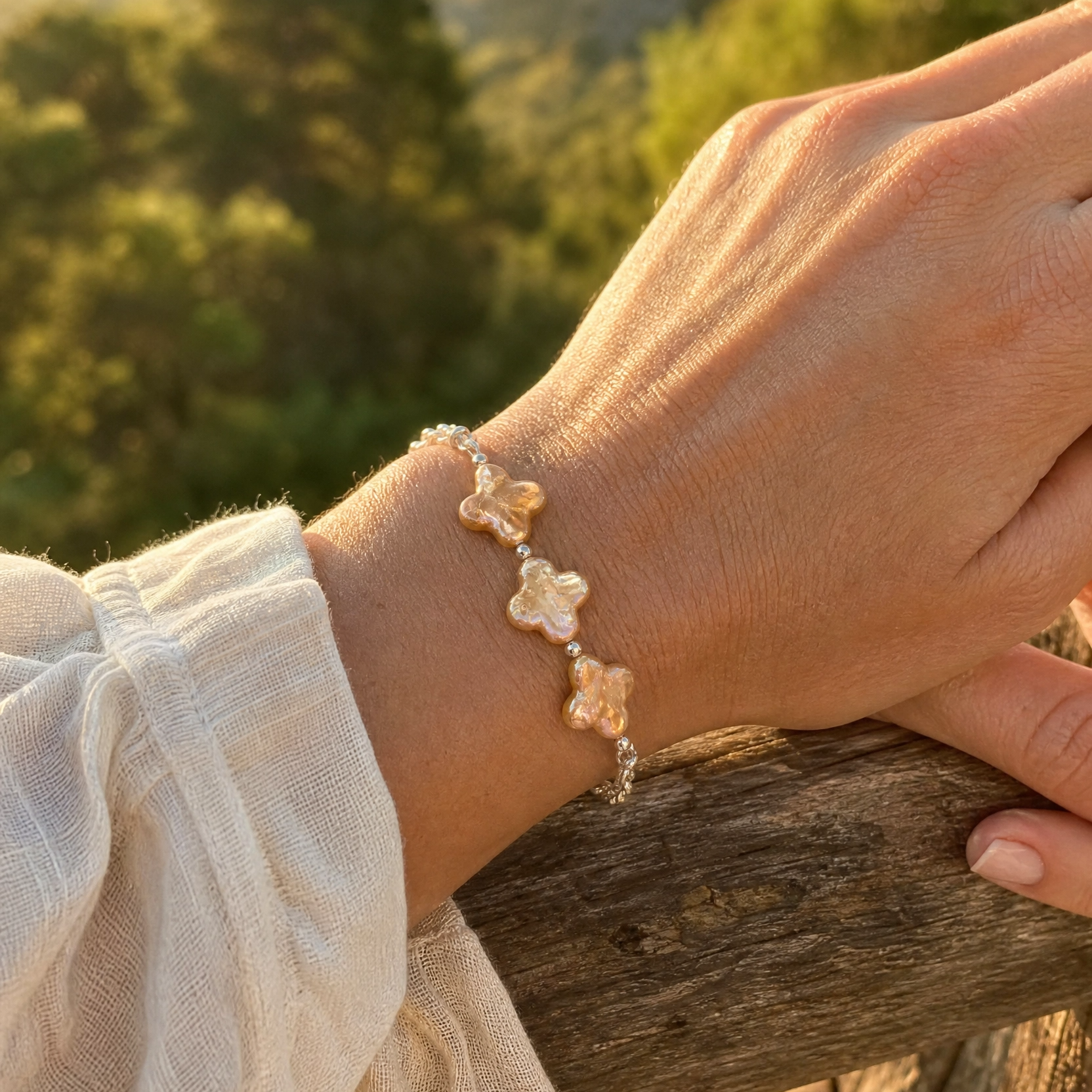 A close-up of a person's wrist wearing a delicate silver rolo chain bracelet with four irregular, champagne-gold tone four leaf clover pearls, set against a blurred outdoor background of green trees and warm sunlight.