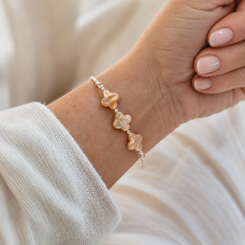 A delicate silver chain bracelet featuring three small, champagne four leaf clover pearls, shown worn on a person's wrist against a soft, warm cream-colored background.