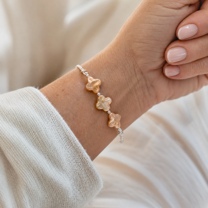 A delicate silver chain bracelet featuring three small, champagne four leaf clover pearls, shown worn on a person's wrist against a soft, warm cream-colored background.