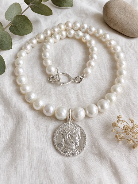 A pearl strand necklace with a silver coin pendant, styled on a textured white fabric with eucalyptus leaves and a stone.