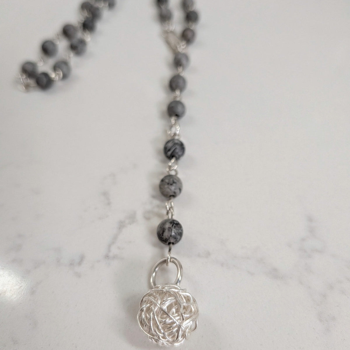 A beaded necklace with dark gray and black marbled picture jasper beads, silver rings, and a silver wire ball pendant, displayed on a white marble surface.