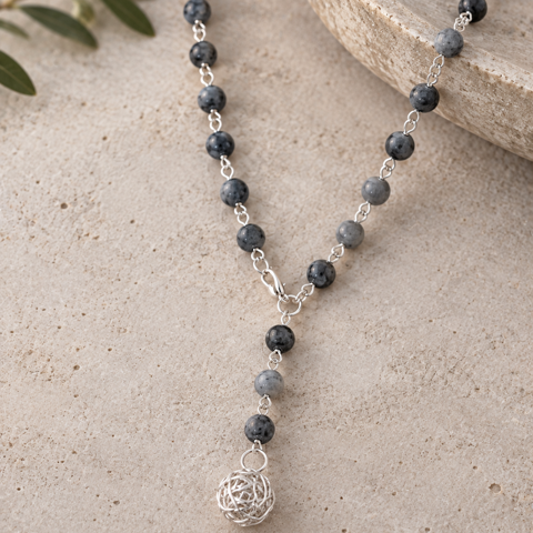 A beaded necklace with dark gray and black round beads, silver links, and a silver wire-wrapped ball pendant, displayed on a textured beige stone surface with a stone dish and olive leaves as props.