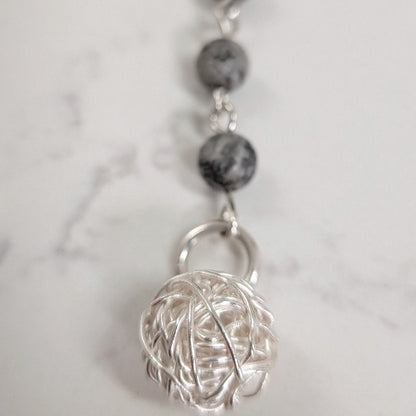A close-up of a dark gray marbled beads and a textured spherical wire ball pendant shown on a light marble-like surface.