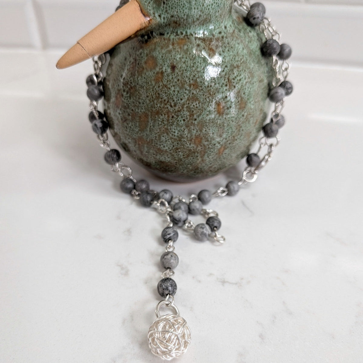 A beaded necklace with gray stone beads, silver and a silver wire ball pendant, draped around a green ceramic duck figurine on a white marble surface.
