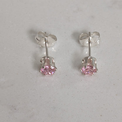 A pair of silver stud earrings with small pink gemstones in a prong setting, displayed on a light gray marble surface.