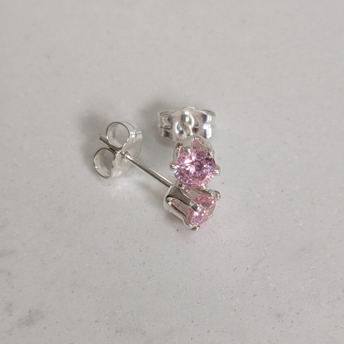 A pair of silver stud earrings with small pink gemstones, shown on a light gray textured surface.