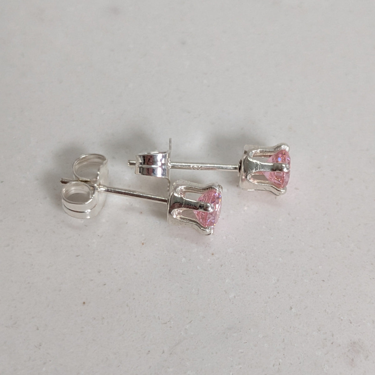 A pair of silver stud earrings with small pink faceted gemstones, displayed on a light gray textured surface.